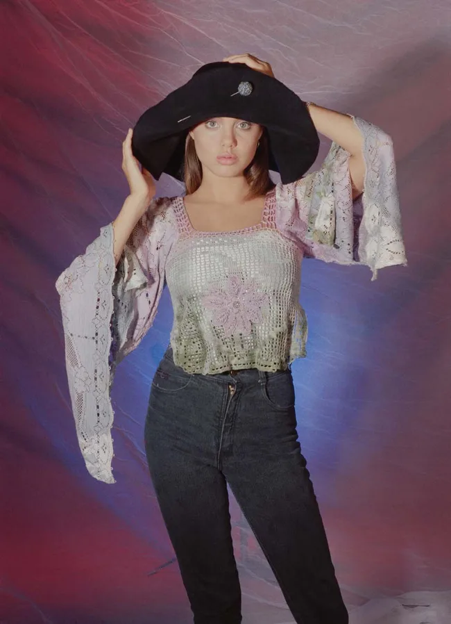A person wearing a black hat and a crochet top against a colorful, abstract background.