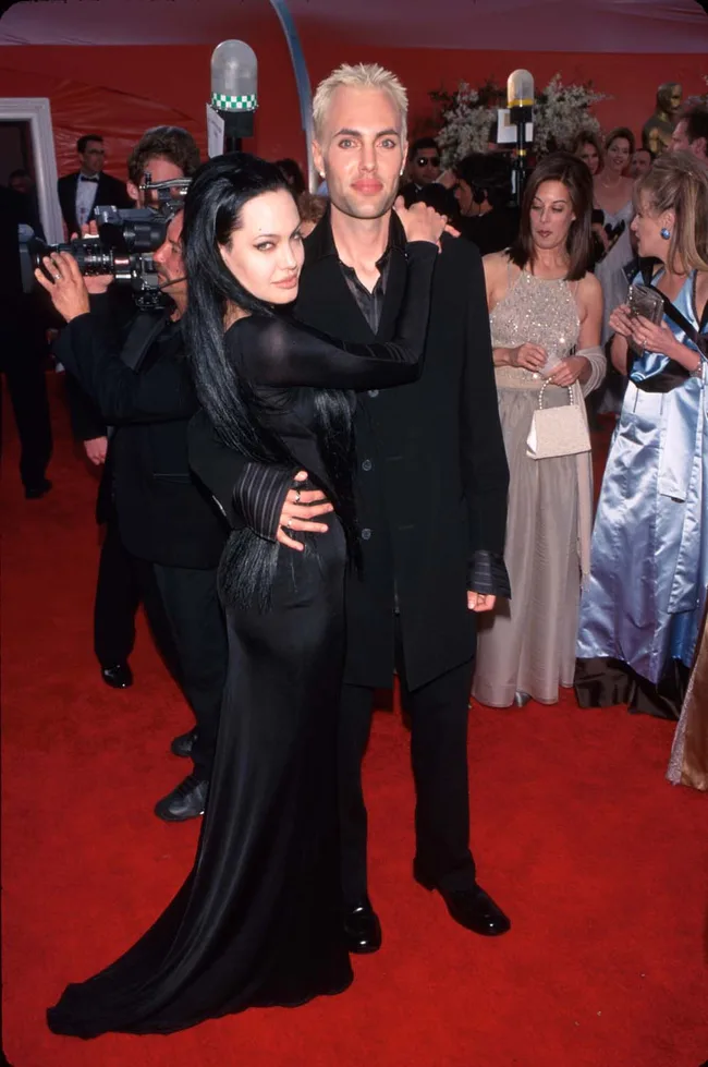 A woman in a black dress embraces a man with blonde hair on a red carpet, surrounded by people and photographers.
