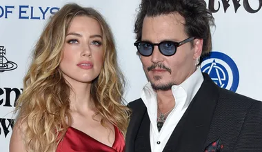 A man in a suit and sunglasses stands beside a woman in a red dress with long hair at a formal event.