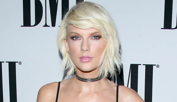 Blonde woman with short hair in front of a BMI logo backdrop, wearing a black choker and dress.