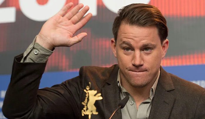 Actor at a press conference, raising his hand, wearing a suit, with a serious expression.