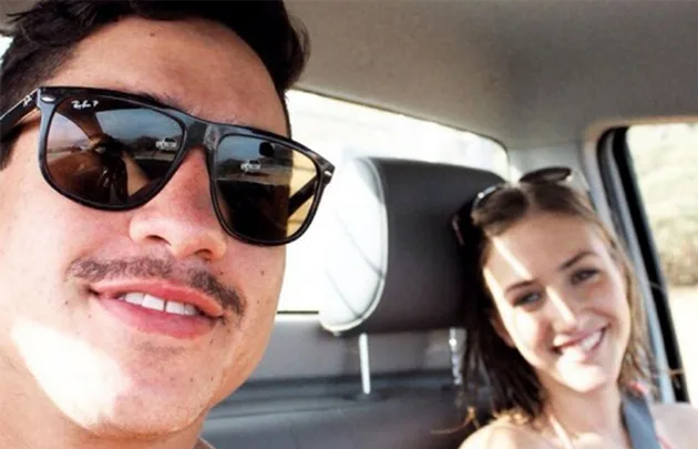 Two people smiling in a car; a man in sunglasses and a woman with long hair.