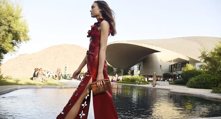 Model in a red dress and holding a brown purse, walking by a reflective pond with modern architecture and mountains in background.
