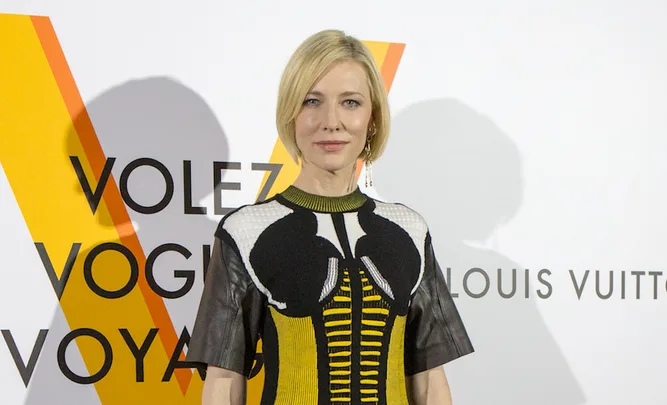 A person in a patterned black and yellow dress stands in front of a backdrop with "Louis Vuitton" text.