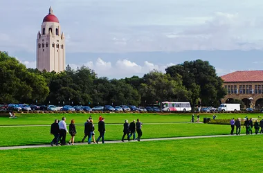 A group of people walks on a path across a green lawn with Hoover Tower in the background at Stanford University.