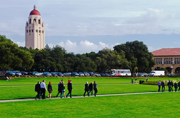 A group of people walks on a path across a green lawn with Hoover Tower in the background at Stanford University.