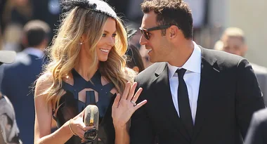 A couple in formal attire smiling at each other outdoors. The woman holds a clutch and wears a black dress with a fascinator.