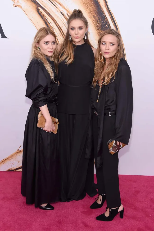 Three women in black outfits pose on a pink carpet in front of a gold and white backdrop.