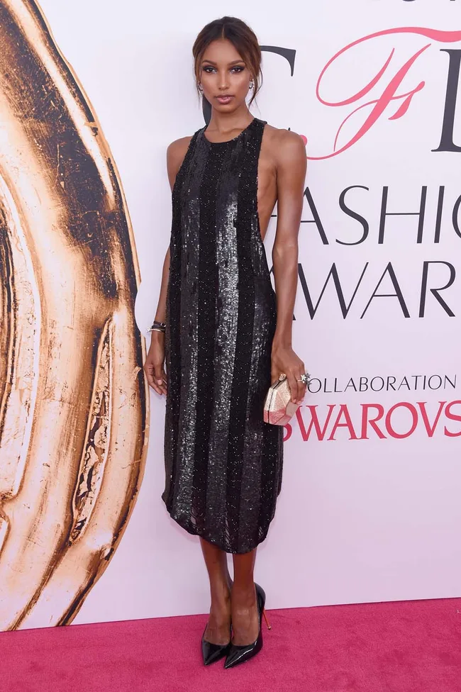 Woman in a black sequined dress on pink carpet at a fashion event, holding a clutch.