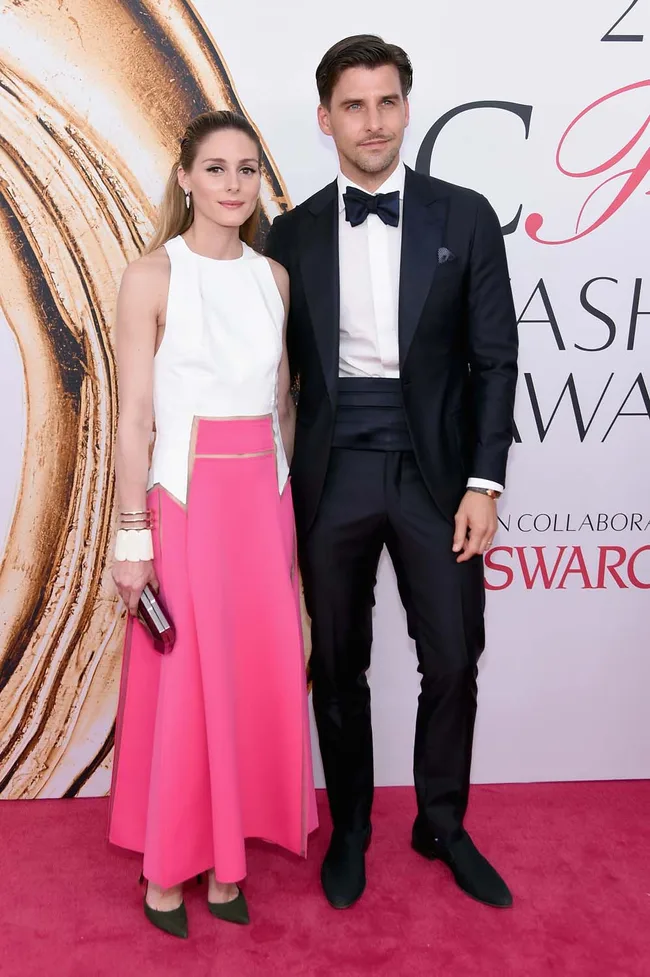 Couple posing at a fashion event, woman in white top and pink skirt, man in black tuxedo with bow tie.