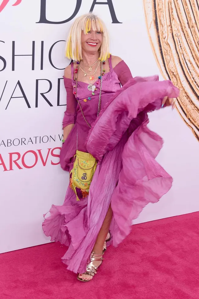 Designer in a vibrant pink dress and colorful accessories poses at a fashion awards red carpet event.