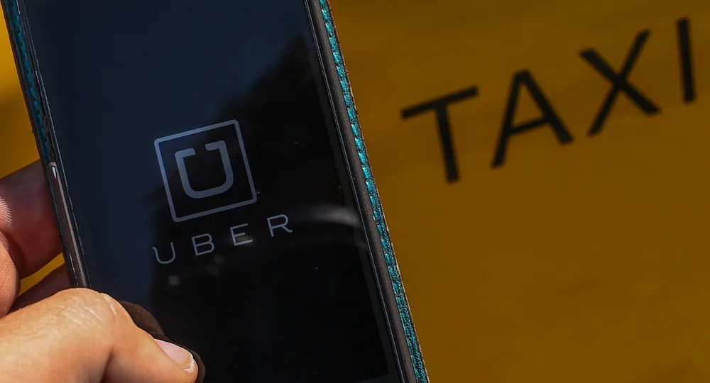Uber app open on a smartphone screen, with a blurred yellow taxi sign in the background.
