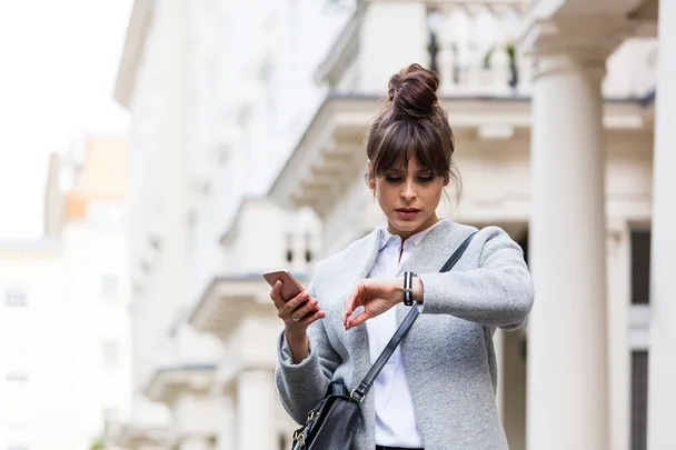 Woman in a gray coat, looking at her watch while holding a smartphone, standing in a city street with white buildings.