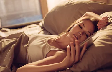 A woman lying in bed, looking at her phone, with warm morning light coming through blinds in the background.