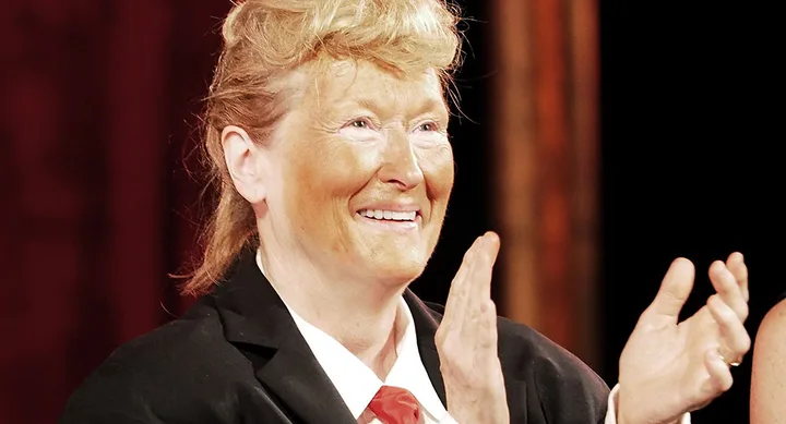 A person with short blond hair and orange makeup claps, wearing a suit and red tie on stage.