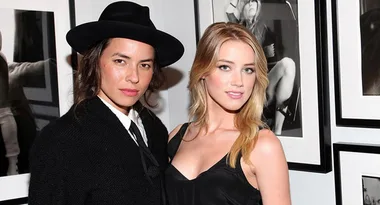 Two people pose together, one wearing a black hat and outfit, the other in a black dress, in front of framed art.