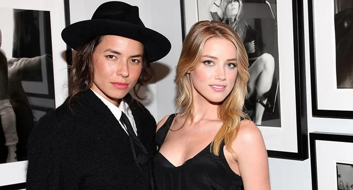 Two people pose together, one wearing a black hat and outfit, the other in a black dress, in front of framed art.