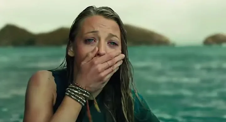 Woman in distress, covering mouth with hand, standing in ocean with blurred hills in the background.