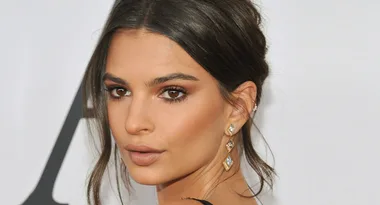 A woman with dark hair tied back, wearing diamond earrings, looks over her shoulder with a neutral expression.