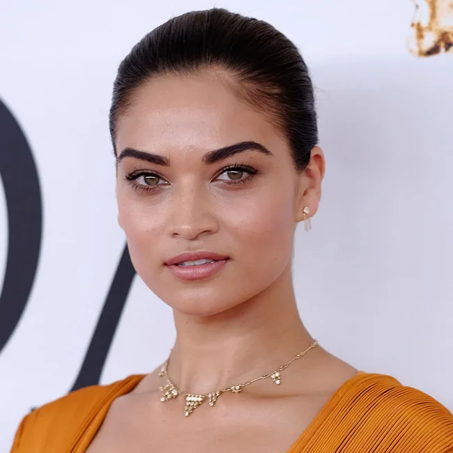 Woman with slicked-back hair wearing an orange dress and gold necklace, posing against a light background.