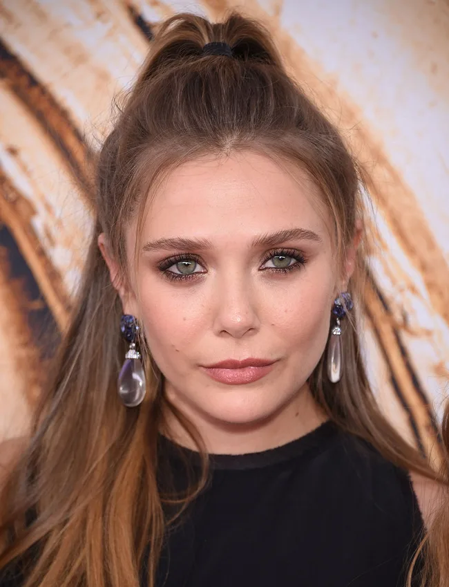 Woman with long brown hair in a ponytail, wearing pearl earrings and a black top against a blurred background.