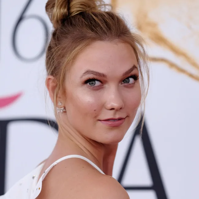 A woman with blonde hair in a bun, wearing a white top, looking over her shoulder, with a blurred background.