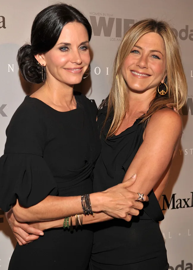 Two women in black dresses embrace and smile on the red carpet at a film event.
