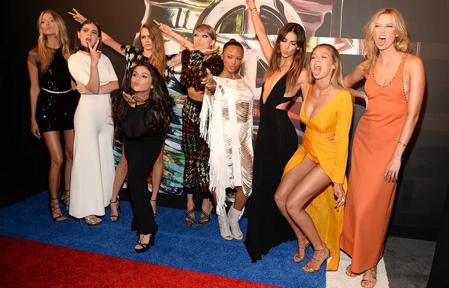 Group of women posing playfully on a red carpet, wearing stylish evening gowns.