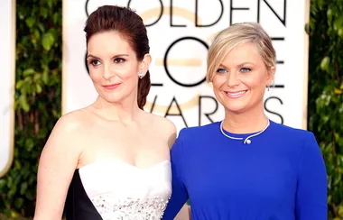 Two women smiling on the red carpet at the Golden Globe Awards, one in a strapless dress and the other in a blue dress.
