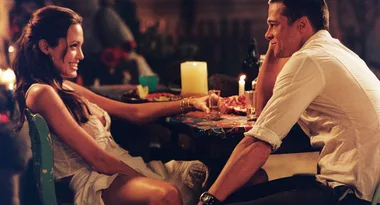 A couple sits across a table, dimly lit by candles, sharing an intimate moment and smiling at each other.