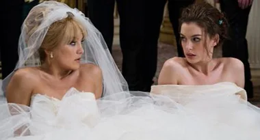 Two brides in wedding dresses sit on the floor, looking surprised at each other.