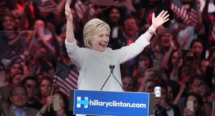A person raises arms and smiles at a podium during a rally, surrounded by cheering crowd with American flags.