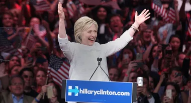 A person raises arms and smiles at a podium during a rally, surrounded by cheering crowd with American flags.