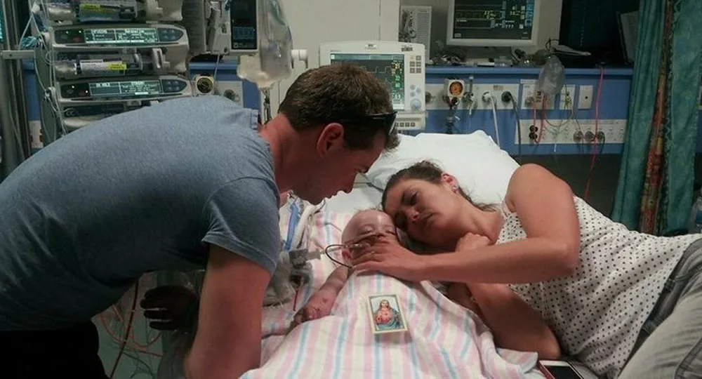 A couple lovingly embraces a baby in a hospital bed surrounded by medical equipment.