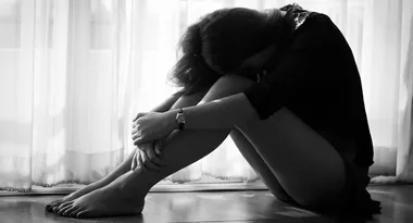 A person sitting on the floor in front of a window, hugging knees, head down, appearing contemplative or sad.