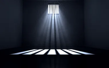 Sunlight streams through a small barred window, casting shadow lines on a dark prison cell floor.