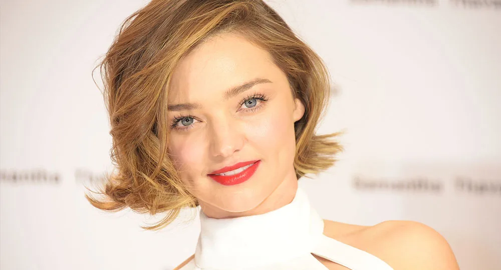 A person with short blonde hair and red lipstick, wearing a white halter top, smiles at the camera.