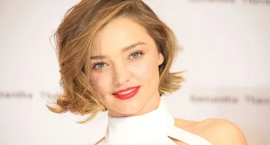 A person with short blonde hair and red lipstick, wearing a white halter top, smiles at the camera.