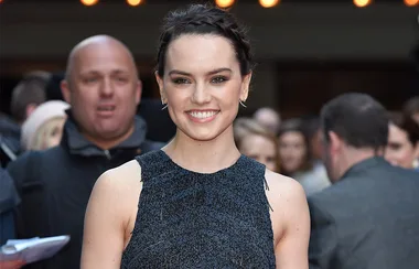 A woman in a dark sleeveless top smiles at a red carpet event, with people in the background.