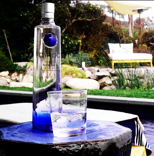 Bottle of Ciroc vodka next to a glass with ice on a table, set in an outdoor garden with chairs in the background.