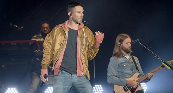 Lead singer in a gold jacket performs on stage with a guitarist, bright stage lights in the background.