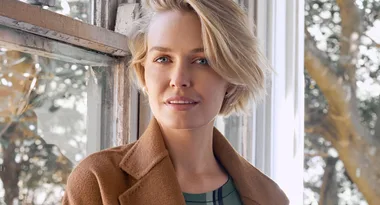 A person with short blonde hair wearing a brown coat stands by a window with a blurred outdoor background.