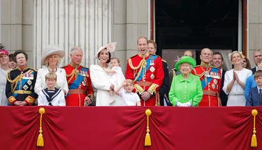 A royal family gathering on a balcony during a formal event, featuring colorful outfits and military uniforms.
