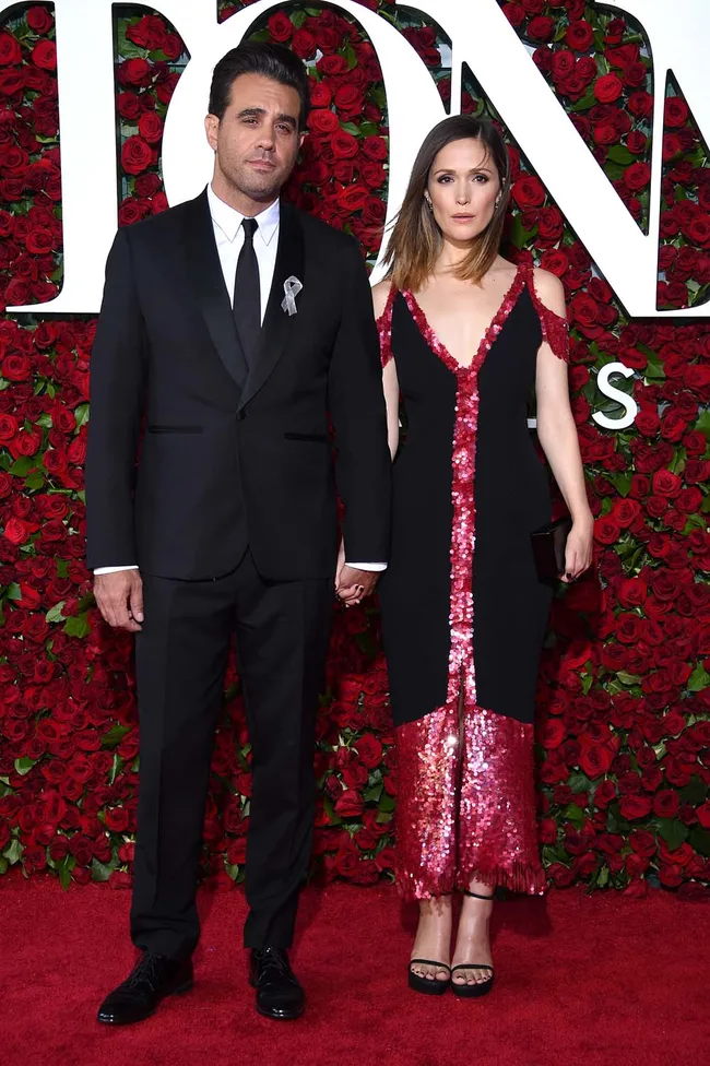 A man in a suit and a woman in a black dress with red sequins hold hands on a red carpet with a rose backdrop.
