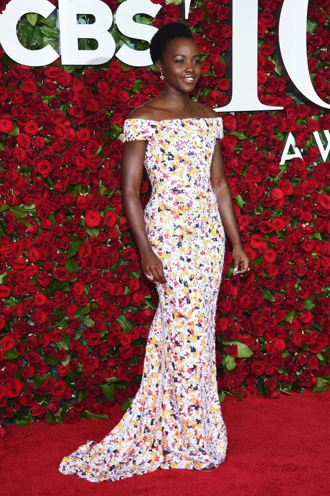 Person in a colorful floral gown on a red carpet, standing against a background of red roses.