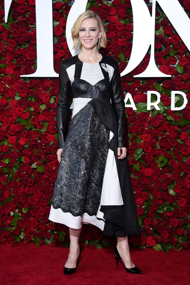 A woman in a black and white dress stands on a red carpet with a rose backdrop at an awards event.