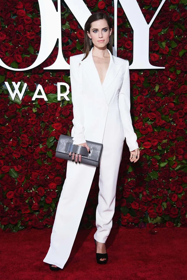 Person in a white outfit on red carpet, holding a gray clutch, with a backdrop of red roses and large text.