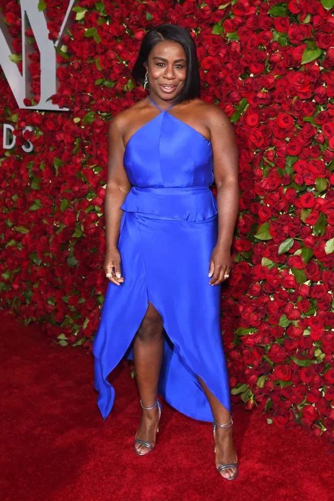 Woman in a vibrant blue dress on a red carpet with a backdrop of red roses.