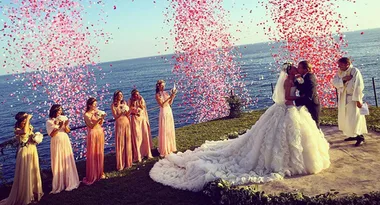 Bride and groom kiss at oceanfront wedding, surrounded by bridesmaids in pink dresses and falling flower petals.