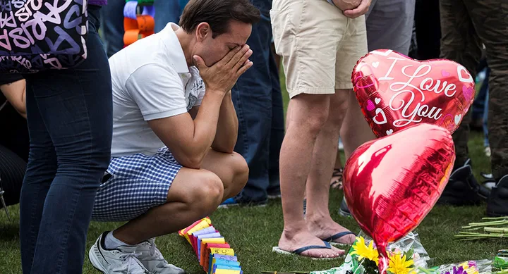 A man kneels at a memorial with "I Love You" heart balloons and flowers, appearing emotional with hands on his face.